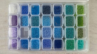 Küçük bölümlerdeki renklere göre sıralanmış tohum boncuklarıyla dolu şeffaf plastik organizatörün üst görünümü. Eşya deposu ve mücevher, tarafsız arkaplanda malzeme üretiyor..