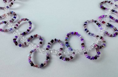 Temiz kristal boncuk zincirine bağlı mor tohum boncuk halkaları ile el yapımı mücevherler. Açık gri zemin üzerinde geniş kopya alanı olan çapraz sınır kompozisyonu. Mücevher tasarımı, el yapımı zanaat ve afiş tasarımları için uygun