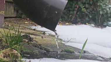 Buz sarkıtı çamurdan kara damlıyor, eriyen buz saçağının yosunlu kaldırıma düşüşünün yakın çekim görüntüsü arka bahçedeki yeşil çimen tarlasının yanında, bulutlu ışık, ince yansıma ve yansıma.,