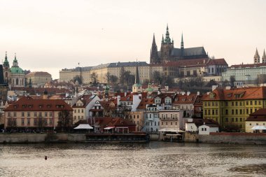 Prague Castle rises above the Vltava River. Along the river there are historical buildings with red roofs. Autumn light casts a soft glow on the stone towers and spiers. March 08, 2026 Prague
