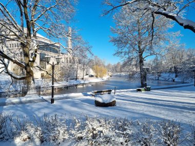 Nehirli güzel bir kış parkı, karla kaplı ağaçlar ve güneşli bir günde modern bir bina. Taze beyaz kar, mavi gökyüzü, uzun gölgeler ve sakin su seyahat, turizm, tatil, doğa için mükemmel huzurlu bir şehir manzarası yaratır.