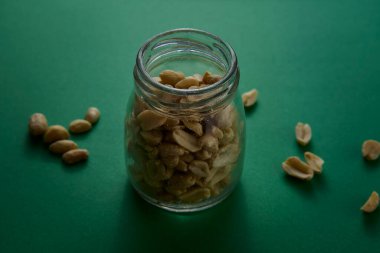 Fıstıkla dolu şeffaf cam bir kavanoz yeşil arka planda duruyor ve etrafa biraz fındık saçılıyor. Minimalist stüdyo hala dokuya ve renge önem veriyor..