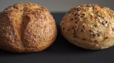 Koyu arkaplana tohum konmuş iki tane fırınlanmış çoklu tahıllı ekmek rulosu. Pastane görselleri için ayrıntılı kabuk ve zanaatkar gıda kavramını gösteren Stüdyo makro döngü videosu.