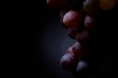 Kırmızı üzümlerin, yumuşak stüdyo ışıklarıyla karanlık bir arkaplanda asılı durduğu yakın plan. Dokuları, tazeliği ve doğal güzelliği vurgulayan sanatsal yemek fotoğrafçılığı kavramı.