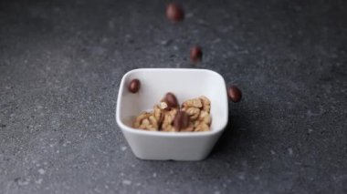 Ceviz ve çikolata kaplı fındıkların koyu gri yüzeyde küçük beyaz bir seramik kaseye düşüşünün yavaş çekim videosu. Yumuşak ışık ve kopyalama alanı ile tatlı ve doğal elementlerin güzel kombinasyonu.