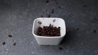 Kavrulmuş kahve çekirdeklerinin gri dokulu bir masada küçük beyaz bir kaseye düşüşünün makro görüntülerini kapatın. Sahne, yeni kavrulmuş kahvenin aromasını, dokusunu ve sıcaklığını ön plana çıkarıyor..