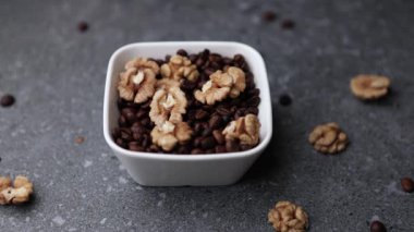 Koyu gri yüzeyde, ceviz ve kavrulmuş kahve çekirdekleri ile dolu küçük beyaz bir seramik kâse etrafında kayan pürüzsüz bir kamera. En düşük ürün bileşimi reklam veya arka plan için idealdir.