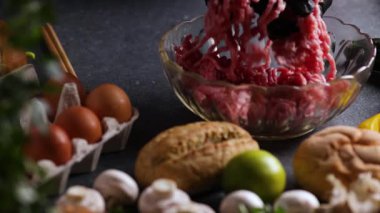 Şefin ellerini mantarların, limonun, çöreklerin, maydanozların ve yumurtaların arasına yerleştirilmiş kristal bir kaseye kıyma koyarken gösteren bir kamera videosu..