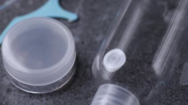 Plastik malzemeler ve hijyen ürünlerini ön plana çıkaran, gri taşlı arka planda diş ipiyle düzenlenmiş, berrak seyahat şişelerinin yavaş bir görüntüsü.