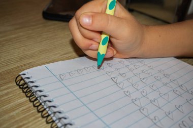 Kalem tutan bir çocuğun eli, çizilmiş bir kağıda kupa şablonu yazıyor, spiral bir deftere çizim yapıyor.