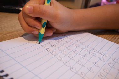 Kalem tutan çocuk, defter sayfasına tekrar tekrar kalp şekilleri çizen, aşkı ve öğrenmeyi simgeleyen.