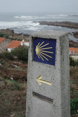 Sarı bir ok ve Camino de Santiago işareti olan taştan bir tabela. Bulutlu havada arkada okyanus ve kayalık bir kıyı var. Yüksek kalite fotoğraf