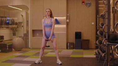 Modern bir fitness merkezinde bir dambılla sumo çömelmesi yapan kararlı genç bir kadın, kaslı kalça ve kalça kasları için daha düşük vücut gücü eğitimine odaklanarak düzgün bir şekilde formda kalmaya çalışıyor..