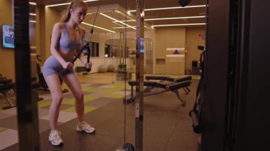Genç, kaslı bir kadın, kablo makinesinin üzerinde triseps egzersizi yapıyor. Ağır çekimde, modern bir fitness merkezinde kas gelişimi için yoğun bir çalışma yapıyor..