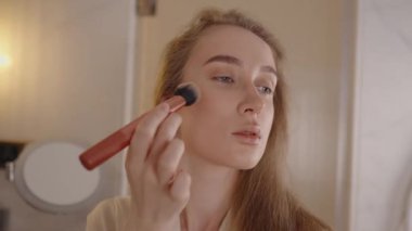 Uzun saçlı, güzel beyaz bir kadın makyaj fırçasıyla yüz pudrası uyguluyor, sabah güneşinin güzelliği sırasında aynaya bakıyor, ılık, aydınlık bir banyoda..