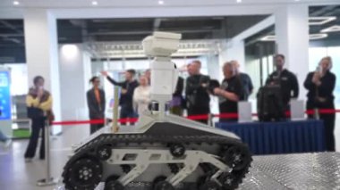 Tırtıl izlerine sahip küçük bir keşif robotu dik bir metal rampaya tırmanarak bir gösteri gerçekleştirirken, mühendisler ve izleyicilerden oluşan bir kalabalık arka planda, robot yeniliği ai.