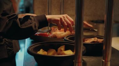 Sıcak büfeden alınmış et ve sebzeleri bir restorandaki bir tabağa koymak için maşa kullanan bir kadının ellerini kapatın. Geleneksel pazar yemeklerinin tadını çıkarın..