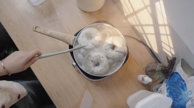 Elektrikli ocağın üzerinde kaynayan bir su kabının içinde tohum ile üç çiğ ev yapımı simit kaynatan bir insanın yavaş çekim görüntüsü, iki silikon spatulayla çevrilmiş..