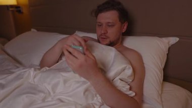 Gece geç saatlerde cep telefonuyla kayarken göz yorgunluğu ve baş ağrısı çeken yorgun bir genç adam uykusuzluk yüzünden uyuyamıyor.