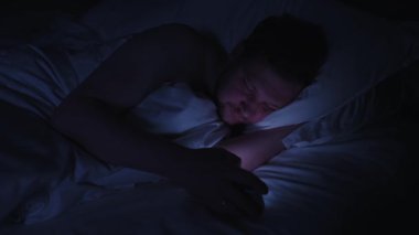Uykusuz bir adam geceleri yatağında cep telefonuna sarılıyor. Ekran ışığı yüzünü karanlık yatak odasında aydınlatıyor. Bağımlılık ve uykusuzluk sorunları yaşıyor.