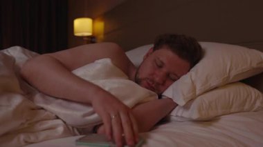 Uykulu adam sabah akıllı telefonuyla çalar saati kapatıyor, gözlerini ovalıyor ve otel odasındaki yumuşak yastıklarla sıcak yorganın altında rahat yatağında uyumaya devam ediyor.
