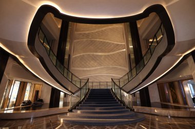 Büyük çok katlı otel lobisi, geniş kıvrımlı merdiven, cam parmaklıklar ve sanat deco duvarlı detaylar, çağdaş bir binada davetkar ve lüks bir atmosfer yaratıyor.