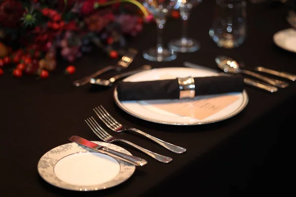 Hassas çatal bıçak takımları, beyaz tabaklar ve siyah peçetelerle hazırlanmış resmi yemek masası, özel bir durum veya kutlama toplantısı için lüks bir ortam yaratıyor.