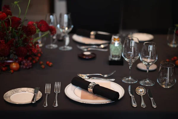 Zarif bir restoran masası, el değmemiş beyaz tabaklar, parlatılmış gümüş takımlar, kristal bardaklar, siyah peçeteler ve canlı kırmızı bir çiçek süsü ile güzel bir şekilde düzenlenmiş.