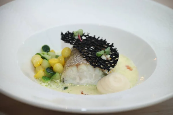 Balık filetosu beyaz bir tabakta sotelenmiş sebzelerle süslenmiş, siyah dekoratif cips ve mikroyeşilliklerle süslenmiş, ince yemek yemekleri sergileniyor.