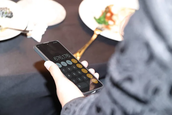 Hesap makinesi açık olan bir akıllı telefonu elinde tutan, tabak ve çatal bıçaklı bir restoranın harcamalarını hesaplayan, finansal planlama ve bütçe hesaplamalarına odaklanan bir kişi.