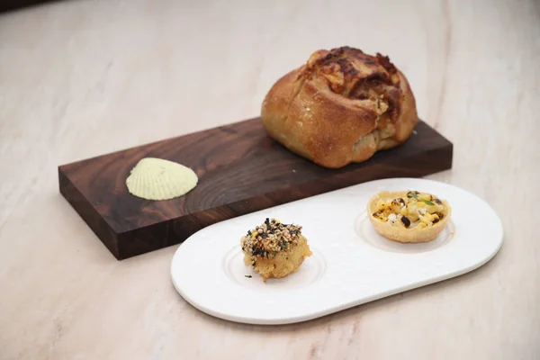 Taze pişmiş ekmek iki rafine edilmiş lokma büyüklüğünde aperatifle servis edilir. Susamlı pasta, ahşap bir tahtada özenle sunulan küçük kiş ve zarif bir yemek deneyimi için beyaz tabak.