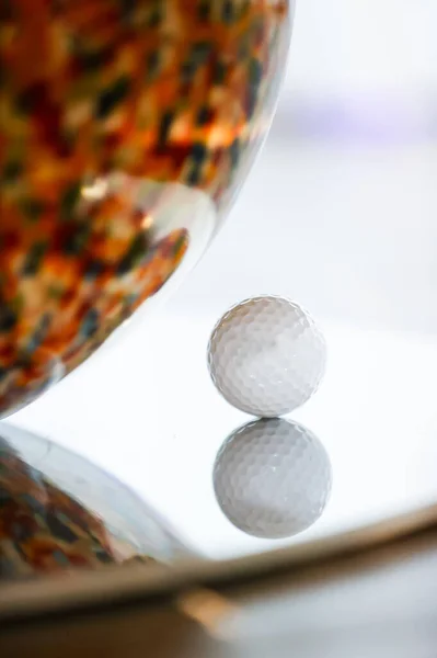 Yansıtıcı yüzeyde duran golf topu, gamzeli dokusunu yansıtıyor, parlak arkaplanda parlak turuncu ve kahverengi benekleri gösteren bulanık cam heykelin yanına yerleştirilmiş golf topu.