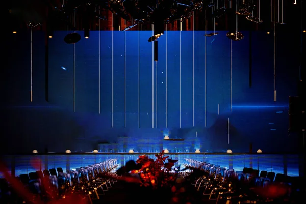 Geniş bir sahne ekranı olan modern balo salonu. Soyut mavi ışıklar, zarif tavan dekorları ve sofistike bir gala ya da kurumsal bir akşam yemeği için hazırlanmış boş masa ve sandalyeler.