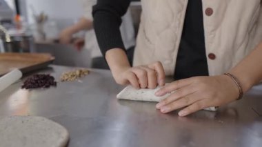 Kadın fırıncı, profesyonel bir fırında, kurutulmuş meyve ve fındıkla dolu taze bir hamur parçasını paslanmaz çelik bir tezgahta dikkatlice yuvarlıyor ve özel bir ekmek hazırlıyor.