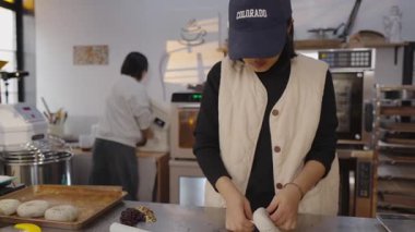 Paslanmaz çelik tezgahın üzerine ustalıkla kep doldurup hamuru saran bir kadın fırıncı profesyonel bir fırında doldurulmuş taze çöreklerle arka planda başka bir meslektaşını hazırlıyor.