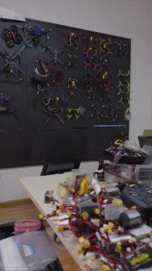 İnsansız hava aracı atölyesindeki dağınık tezgahın yavaş çekim dikey tavası, duvardaki fpv kuadkopterlerinin koleksiyonunu, uzaktan kumandaları, fpv dronları için lityum polimer pilleri, hava araçları