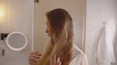 Uzun sarı saçları olan güzel bir kadın. Parlak, modern bir banyoda elleriyle saçlarını tarıyor. Sabah güzellik rutininin tadını çıkarıyor..