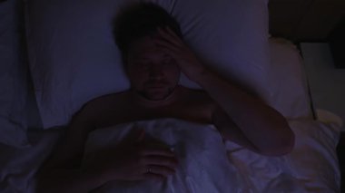 Endişeli adam, uykusuzluk ve endişeyle, uykusuzluktan kıvranıyor, stres yüzünden uyuyamıyor, alnına dokunuyor ve karanlık yatak odasında huzursuz yatıyor, uyuyamıyor.