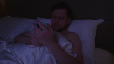 Yoğunlaşmış genç adam gece geç saatte yatakta yatarken akıllı telefonunu kaydırıyor ve aşırıyor. Uyumadan önce karanlık bir yatak odasında uykusuzluk ve ekran bağımlılığından muzdarip.
