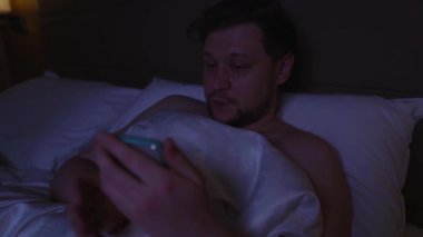 Genç adam gece yatakta uyanık yatıyor, akıllı telefonunu karanlıkta kaydırıyor, uykusuzluk çekiyor, uyuyamıyor, uyku düzenini bozuyor, zihinsel sağlığını etkileyen ekran ve dinlenmeyi engelliyor.