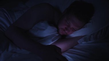 Telefon bağımlılığı olan uykusuz bir adam geceleri karanlık bir yatak odasında sosyal medyada geziniyor. Parlak ekran ışığı ve mavi ışıkta uykusuzlukla boğuşuyor.
