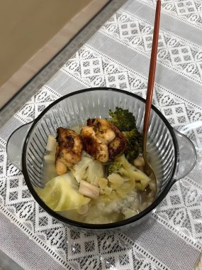 Süslü bir masa örtüsünün üzerinde cam bir kasede servis edilen tavuk ve sebzeli sağlıklı ve berrak çorba.