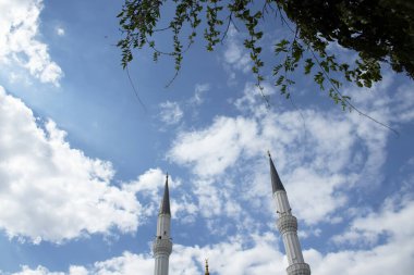 Bulutlu mavi gökyüzünün altında yeşil yapraklarla çerçevelenmiş iki cami minaresi. Doğa ve İslami mimariyi barışçıl kompozisyonda birleştirir. Seyahat ve manevi görsellik için ideal..