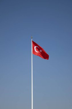 Red Turkish flag with crescent and star waving on a tall flagpole against a clean blue sky. Perfect for stories on Turkey, patriotism, national days, travel, politics, news design, or minimalist backgrounds.