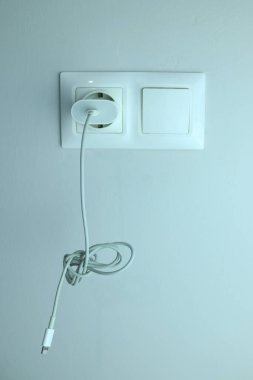 White charging cable plugged into modern wall socket on light wall. Minimalistic photo symbolizing technology, power, electricity, and home connection.