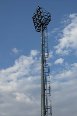 Mavi gökyüzüne uzanan kafesli merdiveni olan uzun metal projektör kulesi, detaylı alçak açılı görüş, spor tesisleri, mühendislik, güvenlik, altyapı, modelleme, kopyalama alanı için ışıklandırma ekipmanları ve yapıyı gösteriyor..