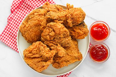 Kırmızı ve beyaz kareli kumaşın üzerinde yuvarlak beyaz bir tabakta kızarmış tavuk servisinin üst görüntüsü, mermer bir yüzeye iki küçük cam kase kırmızı sos ile eşleştirilmiş.