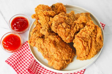 Kırmızı ve beyaz kareli kumaşın üzerinde yuvarlak beyaz bir tabak üzerinde kızarmış tavuk servisi, mermer bir yüzey üzerinde iki küçük cam kase kırmızı sosla eşleştirilmiş.