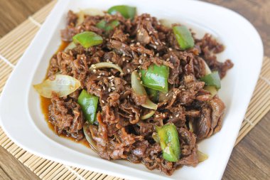 Yeşil çan biberi ve soğanla kızartılmış ve üzerine beyaz susam tohumları serpiştirilmiş yumuşak marine edilmiş sığır eti dilimleri. Tabak, açık bambu bir hasırın üzerinde beyaz dikdörtgen bir tabakta sunuluyor..