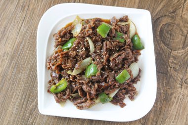 Salamura edilmiş sığır eti dilimleri, yeşil dolmalık biber ve soğanla kızartılmış, üzerine beyaz susam tohumları serpiştirilmiş. Tabak, ahşap bir arka planda beyaz dikdörtgen bir tabakta sunuluyor..